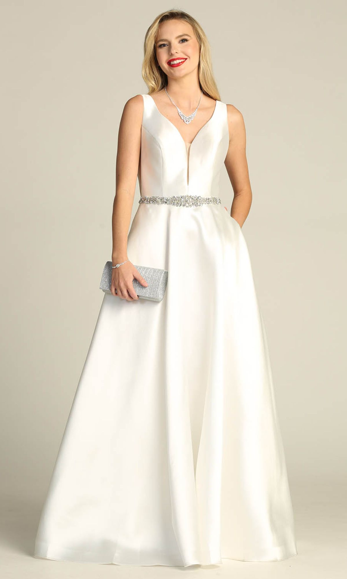 Woman in the Jeweled-Waist Long Prom Ball Gown: Chicas CC3021, a sleeveless white satin A-line dress with deep V-neckline and jeweled waist belt, styled with a silver necklace and clutch, stands against a plain background.